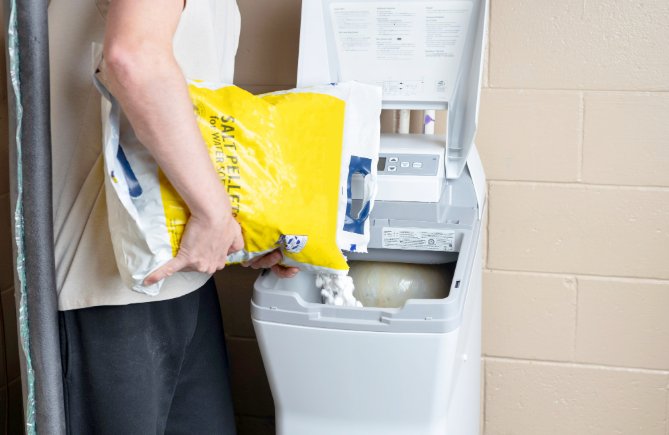 A person pours salt pellets from a yellow bag into the open compartment of a water softener in a utility room, highlighting Why Your Bedfordshire Home Needs a Water Softener to ensure consistently soft and clean water throughout the house.
